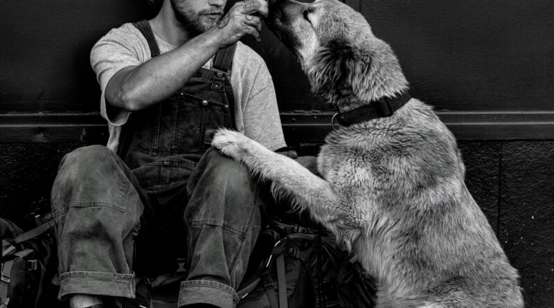 man in black t shirt and black pants sitting on floor with dog in grayscale photography