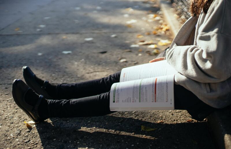 crop faceless student with textbook sitting on ground in park