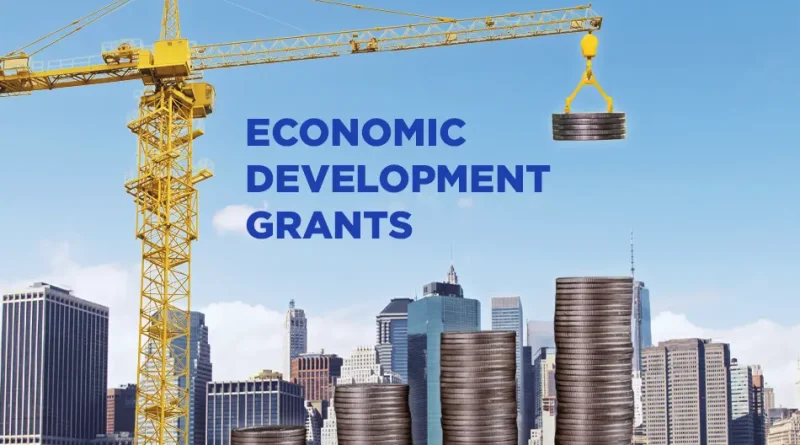 A large yellow construction crane lifts a stack of coins above several tall piles of coins against a city skyline. Blue text reads, “Check out these Economic Development Grants,” with a blue arrow pointing right. The GrantWatch logo appears at the bottom.