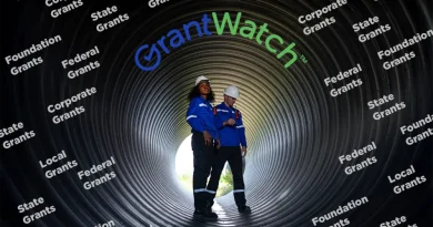 Two workers wearing safety helmets and blue uniforms stand inside a large industrial pipe. The word “GrantWatch” appears above them, surrounded by repeated text labels such as “Federal Grants,” “State Grants,” “Local Grants,” “Corporate Grants,” and “Foundation Grants,” representing different types of funding sources.