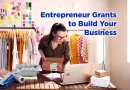 A young female entrepreneur wearing glasses works on her laptop in a bright studio filled with clothing and shipping boxes. Text on the image reads, “Entrepreneur Grants to Build Your Business.”