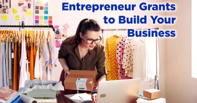 A young female entrepreneur wearing glasses works on her laptop in a bright studio filled with clothing and shipping boxes. Text on the image reads, “Entrepreneur Grants to Build Your Business.”