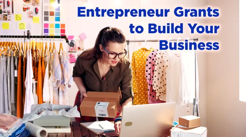 A young female entrepreneur wearing glasses works on her laptop in a bright studio filled with clothing and shipping boxes. Text on the image reads, “Entrepreneur Grants to Build Your Business.”