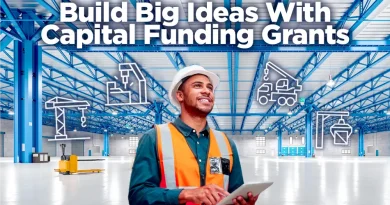 A construction project manager wearing a hard hat and safety vest smiles while holding a tablet inside a large, bright industrial warehouse. Blue steel beams span the ceiling, and white line-art icons of construction equipment—cranes, drills, and trucks—float around him. Bold text reads: 'Build Big Ideas With Capital Funding Grants.'