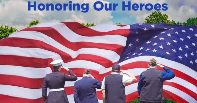 Four veterans stand on a grassy field saluting a large American flag that fills the background, with the words “Honoring Our Heroes” written above them against a partly cloudy sky.