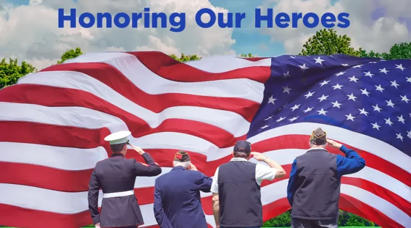Four veterans stand on a grassy field saluting a large American flag that fills the background, with the words “Honoring Our Heroes” written above them against a partly cloudy sky.