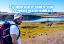 A smiling hiker wearing a hat and backpack stands near a river surrounded by grasses, elk, and waterfowl, with the text “Funding and In-Kind Grants to Protect Water Habitats” displayed over the landscape.