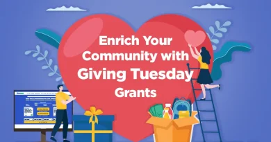 Illustration of two people placing hearts and gifts in front of a large red heart with the text “Enrich Your Community with Giving Tuesday Grants.” One person stands on a ladder holding a heart, while another stands near a computer displaying the GrantWatch website. A gift box and donation items sit at the center. The GrantWatch logo is at the bottom right.