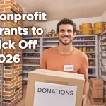 Smiling volunteer holding a donation box inside a nonprofit food pantry, surrounded by shelves of food, with text reading “Nonprofit Grants to Kick Off 2026.”
