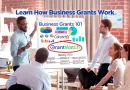 Group of professionals in a meeting room gathered around a flip chart titled “Learn How Business Grants Work.” The chart reads “Business Grants 101” and displays colorful charts, a business plan diagram, and the GrantWatch logo circled in red, as one person presents and others listen attentively.