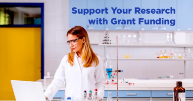 A woman wearing safety glasses and a white lab coat works on a laptop in a modern laboratory, surrounded by glass beakers, test tubes, and scientific equipment, with the text “Support Your Research with Grant Funding” displayed above her.