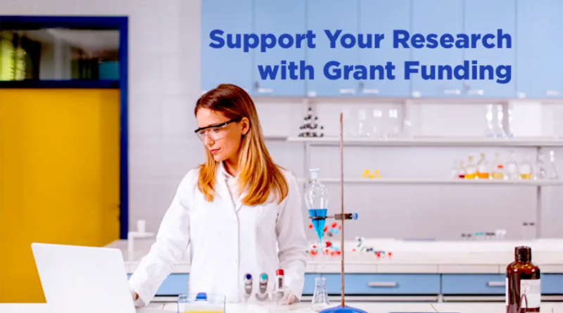 A woman wearing safety glasses and a white lab coat works on a laptop in a modern laboratory, surrounded by glass beakers, test tubes, and scientific equipment, with the text “Support Your Research with Grant Funding” displayed above her.