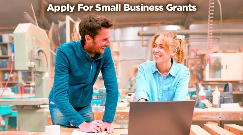 Two small business owners smiling and talking in a workshop while working at a table with a laptop and woodworking tools. Text on the image reads “Apply For Small Business Grants.”