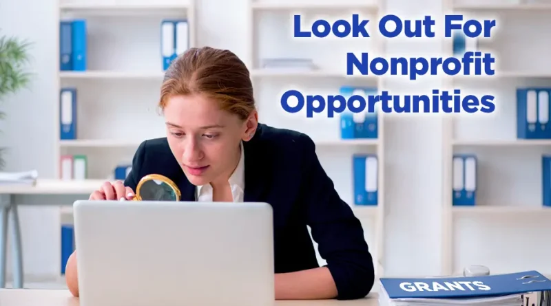 Woman in business attire examines a laptop screen with a magnifying glass at a desk; shelves of binders behind her and a folder labeled “GRANTS” nearby. Text reads, “Look Out For Nonprofit Opportunities.”