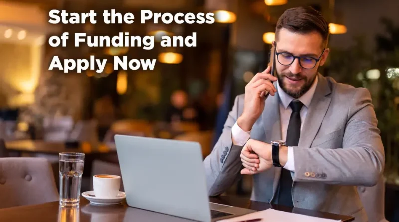 A well-dressed man in a suit sits at a table in a warmly lit café, working on a laptop while talking on his phone and checking his watch. On the table are a cup of coffee and a glass of water. Large text on the left side reads: “Start the Process of Funding and Apply Now.”