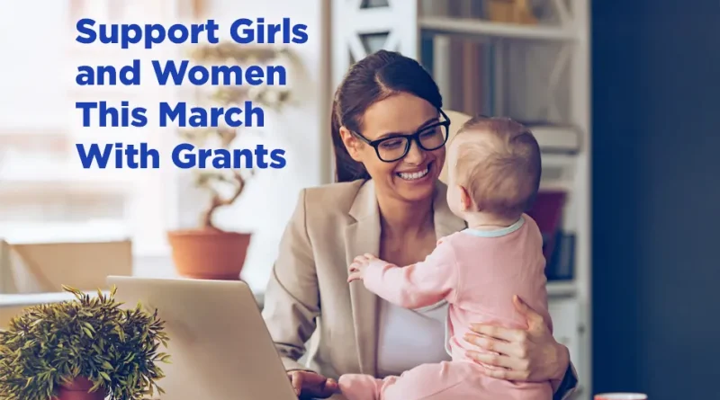 Support Girls and Women This March With Grants. A smiling woman wearing glasses holds a baby on her lap while working on a laptop at a desk, with a plant and coffee mug nearby in a bright home office setting.