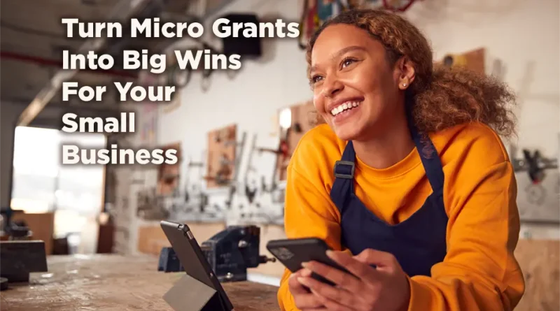 A smiling small business owner in a workshop leans on a wooden workbench while holding a phone, with tools and equipment blurred in the background. Overlaid text reads: “Turn Micro Grants Into Big Wins For Your Small Business.”