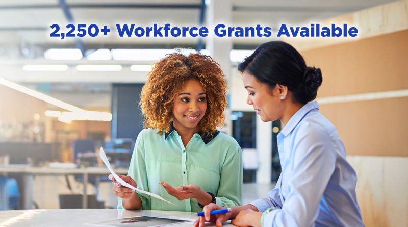 Two professional women reviewing documents together in a modern office setting. One woman with curly hair holds a paper and gestures while speaking, as the other woman listens and holds a pen. Large text on the left side of the image reads, “Fast Track Your Business Career With Workforce Grants.”