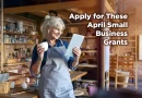Smiling small business owner in a workshop apron holding a tablet and coffee mug, standing in a creative studio space, with text overlay reading “Apply for These April Small Business Grants.”