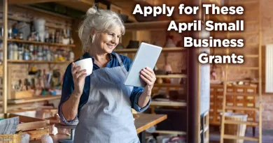 Smiling small business owner in a workshop apron holding a tablet and coffee mug, standing in a creative studio space, with text overlay reading “Apply for These April Small Business Grants.”
