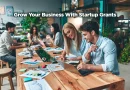 A group of people working on separate projects. The focus is on the two people at the end of a long wooden table. The room gives off an organic vibe with wood, plants, and smiling people. "Grow Your Business with Startup Grants."