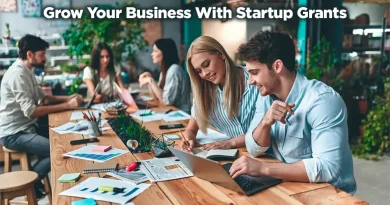 A group of people working on separate projects. The focus is on the two people at the end of a long wooden table. The room gives off an organic vibe with wood, plants, and smiling people. "Grow Your Business with Startup Grants."
