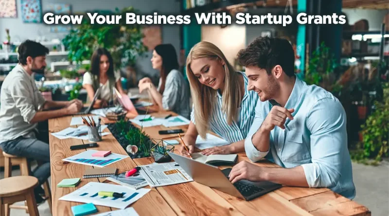 A group of people working on separate projects. The focus is on the two people at the end of a long wooden table. The room gives off an organic vibe with wood, plants, and smiling people. "Grow Your Business with Startup Grants."