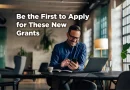 Man sits at his laptop but typing on his phone and smiling. The caption reads: Be the First to Apply for These New Grants