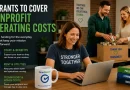 Smiling nonprofit staff members and volunteers work together in an office while packing donation boxes and managing tasks on a laptop. Text on the image reads “5 Grants to Cover Nonprofit Operating Costs” with examples including staff and benefits, rent and utilities, and technology and admin costs. GrantWatch branding appears on a coffee mug beside the computer, while books labeled programs, community, and impact sit nearby.