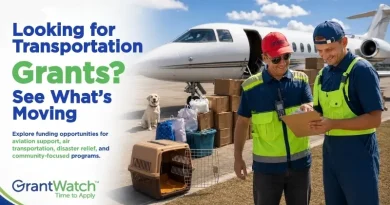 A GrantWatch promotional graphic featuring two transportation workers reviewing a clipboard beside a private jet and stacked cargo boxes on an airport runway. A dog sits near pet carriers and supplies in the background. The text reads, “Looking for Transportation Grants? See What’s Moving,” and highlights funding opportunities for aviation support, air transportation, disaster relief, and community-focused programs. The GrantWatch logo appears in the bottom left corner.