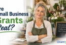 A promotional image for GrantWatch featuring a smiling older woman standing in a flower shop with her arms crossed, wearing a green apron. Large text on the left reads, “Are Small Business Grants Real?” in navy blue and green lettering. A chalkboard sign in the background says, “Your passion. Your purpose. Funding can help it grow.” The GrantWatch logo appears in the bottom right corner.