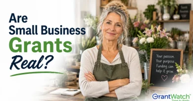 A promotional image for GrantWatch featuring a smiling older woman standing in a flower shop with her arms crossed, wearing a green apron. Large text on the left reads, “Are Small Business Grants Real?” in navy blue and green lettering. A chalkboard sign in the background says, “Your passion. Your purpose. Funding can help it grow.” The GrantWatch logo appears in the bottom right corner.