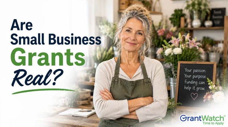 A promotional image for GrantWatch featuring a smiling older woman standing in a flower shop with her arms crossed, wearing a green apron. Large text on the left reads, “Are Small Business Grants Real?” in navy blue and green lettering. A chalkboard sign in the background says, “Your passion. Your purpose. Funding can help it grow.” The GrantWatch logo appears in the bottom right corner.