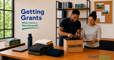 Two nonprofit staff members sort donated clothing into a donation box in an office setting. Text on the wall reads, “Getting Grants When You’re a New Nonprofit.” Folded clothing, a reusable water bottle, books, and a laptop sit on a table. The GrantWatch logo appears in the bottom right corner.