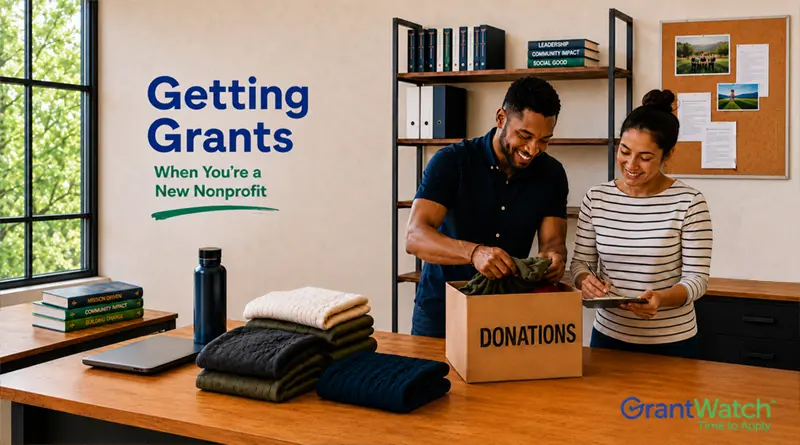 Two nonprofit staff members sort donated clothing into a donation box in an office setting. Text on the wall reads, “Getting Grants When You’re a New Nonprofit.” Folded clothing, a reusable water bottle, books, and a laptop sit on a table. The GrantWatch logo appears in the bottom right corner.