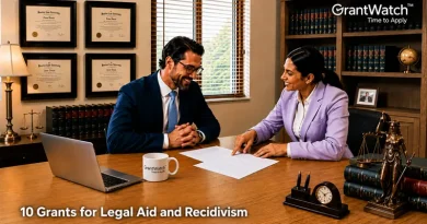 Two legal professionals sit across from each other in a law office, reviewing documents at a wooden desk. One points to paperwork while discussing details with the other. The office features framed law degrees, bookshelves filled with legal volumes, a laptop, and scales of justice decor. Text on the image reads: “10 Grants for Legal Aid and Recidivism.” GrantWatch logo appears in the upper-right corner.