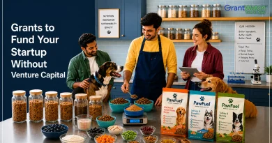 Three people in a pet food startup kitchen and testing lab prepare dog food recipes while interacting with two dogs. Shelves of ingredients, measuring tools, bowls of food, and packaged pet food products are displayed on a worktable. Large text on the left reads, “Grants to Fund Your Startup Without Venture Capital,” with the GrantWatch logo in the upper-right corner.