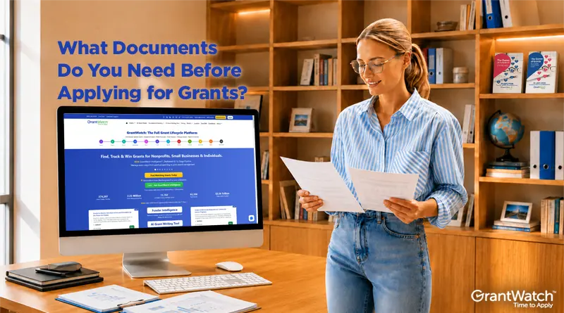 A professional woman stands in a modern office reviewing documents beside a desktop computer displaying the GrantWatch Full Grant Lifecycle Platform. Large text reads, “What Documents Do You Need Before Applying for Grants?” The workspace features bookshelves, binders, a globe, and organized paperwork, highlighting grant preparation, document management, and funding research. GrantWatch logo appears in the lower right corner.