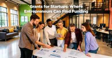 A diverse group of entrepreneurs and startup professionals gather around a table in a modern coworking space, reviewing business plans and funding strategies. Text overlay reads: "Grants for Startups: Where Entrepreneurs Can Find Funding."