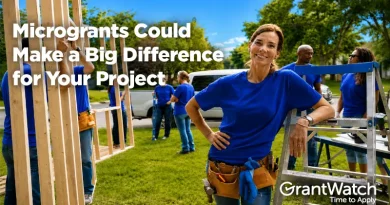 Volunteers wearing blue shirts work together on a community construction project outdoors. A woman in a tool belt stands beside building materials while text reads, “Microgrants Could Make a Big Difference for Your Project.” GrantWatch logo appears in the corner.