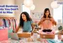 Two women prepare online retail orders in a small boutique. One packs a shipping box while working at a laptop, and the other folds a peach-colored shirt over tissue paper. Clothing racks, branded packaging, shipping supplies, and a workspace filled with stickers and packing materials surround them. Overlay text reads, "5 Small Business Grants You Don't Want to Miss," with the GrantWatch logo in the upper-right corner.