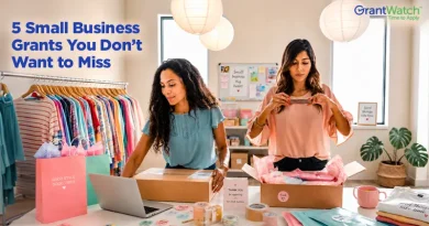 Two women prepare online retail orders in a small boutique. One packs a shipping box while working at a laptop, and the other folds a peach-colored shirt over tissue paper. Clothing racks, branded packaging, shipping supplies, and a workspace filled with stickers and packing materials surround them. Overlay text reads, "5 Small Business Grants You Don't Want to Miss," with the GrantWatch logo in the upper-right corner.
