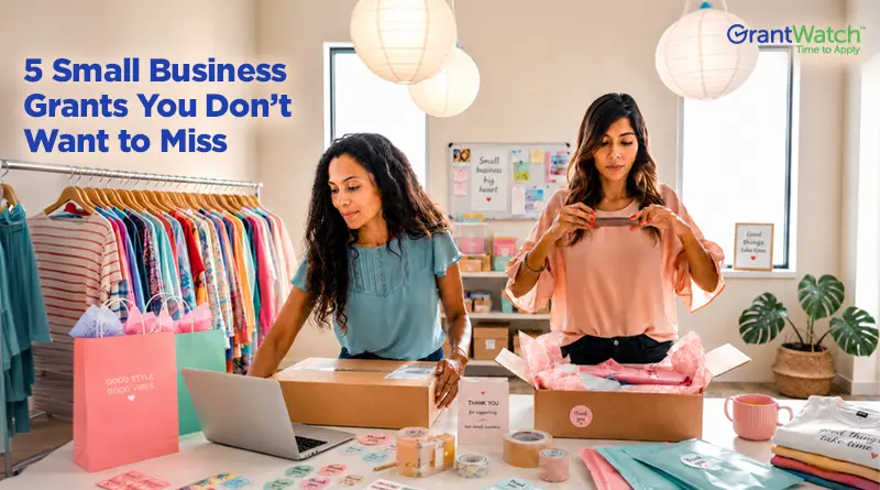 Two women prepare online retail orders in a small boutique. One packs a shipping box while working at a laptop, and the other folds a peach-colored shirt over tissue paper. Clothing racks, branded packaging, shipping supplies, and a workspace filled with stickers and packing materials surround them. Overlay text reads, "5 Small Business Grants You Don't Want to Miss," with the GrantWatch logo in the upper-right corner.
