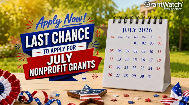 Patriotic-themed GrantWatch graphic promoting July nonprofit grants. A bold red, white, and blue sign reads, "Apply Now! Last Chance to Apply for July Nonprofit Grants." Beside it, a desktop calendar displays July 2026. Red, white, and blue decorations, including ribbons, stars, and small American flag accents, are arranged on a wooden table outdoors with a blurred green background. The GrantWatch logo appears in the top-right corner.