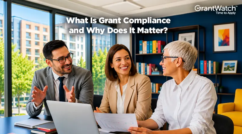 Three professionals sit around a conference table in a modern office, discussing documents and collaborating on a grant-related project. Large windows overlook city buildings, while bookshelves and framed artwork create a professional setting. Text on the image reads, “What Is Grant Compliance and Why Does It Matter?” with the GrantWatch logo displayed in the upper-right corner.