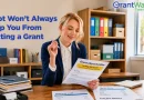 A woman in business attire sits at a desk reviewing a document titled "Grant Opportunities." Office shelves with binders and supplies are visible in the background. Papers on the desk reference business loans, regular costs, and past-due balances. Overlay text reads, "Debt Won’t Always Stop You From Getting a Grant," with the GrantWatch logo displayed in the upper-right corner.