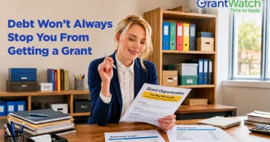 A woman in business attire sits at a desk reviewing a document titled "Grant Opportunities." Office shelves with binders and supplies are visible in the background. Papers on the desk reference business loans, regular costs, and past-due balances. Overlay text reads, "Debt Won’t Always Stop You From Getting a Grant," with the GrantWatch logo displayed in the upper-right corner.