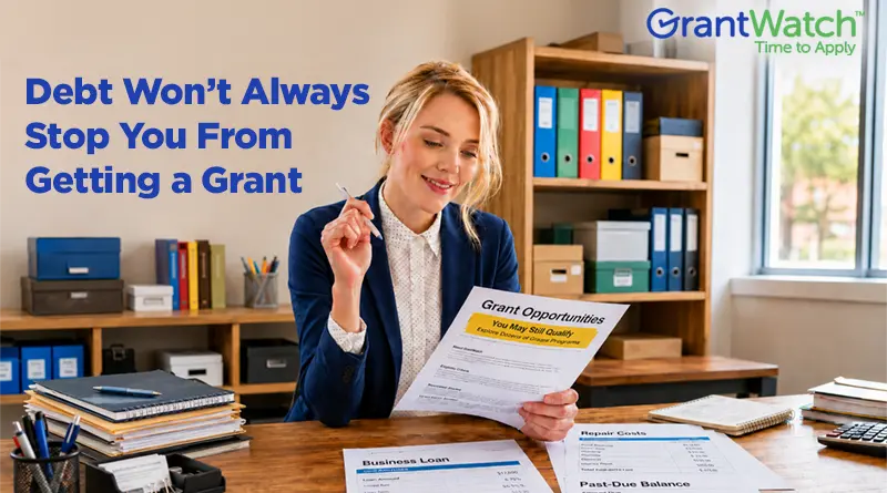 A woman in business attire sits at a desk reviewing a document titled "Grant Opportunities." Office shelves with binders and supplies are visible in the background. Papers on the desk reference business loans, regular costs, and past-due balances. Overlay text reads, "Debt Won’t Always Stop You From Getting a Grant," with the GrantWatch logo displayed in the upper-right corner.
