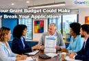 A group of five professionals sits around a conference table discussing a grant budget during a business meeting. One man holds a budget document while others review paperwork and collaborate. The image features the headline, "Your Grant Budget Could Make or Break Your Application," with the GrantWatch logo in the upper-right corner. The modern office includes large windows, colorful wall art, and a sign reading "Strategy, Focus, Impact."