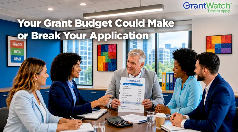 A group of five professionals sits around a conference table discussing a grant budget during a business meeting. One man holds a budget document while others review paperwork and collaborate. The image features the headline, "Your Grant Budget Could Make or Break Your Application," with the GrantWatch logo in the upper-right corner. The modern office includes large windows, colorful wall art, and a sign reading "Strategy, Focus, Impact."